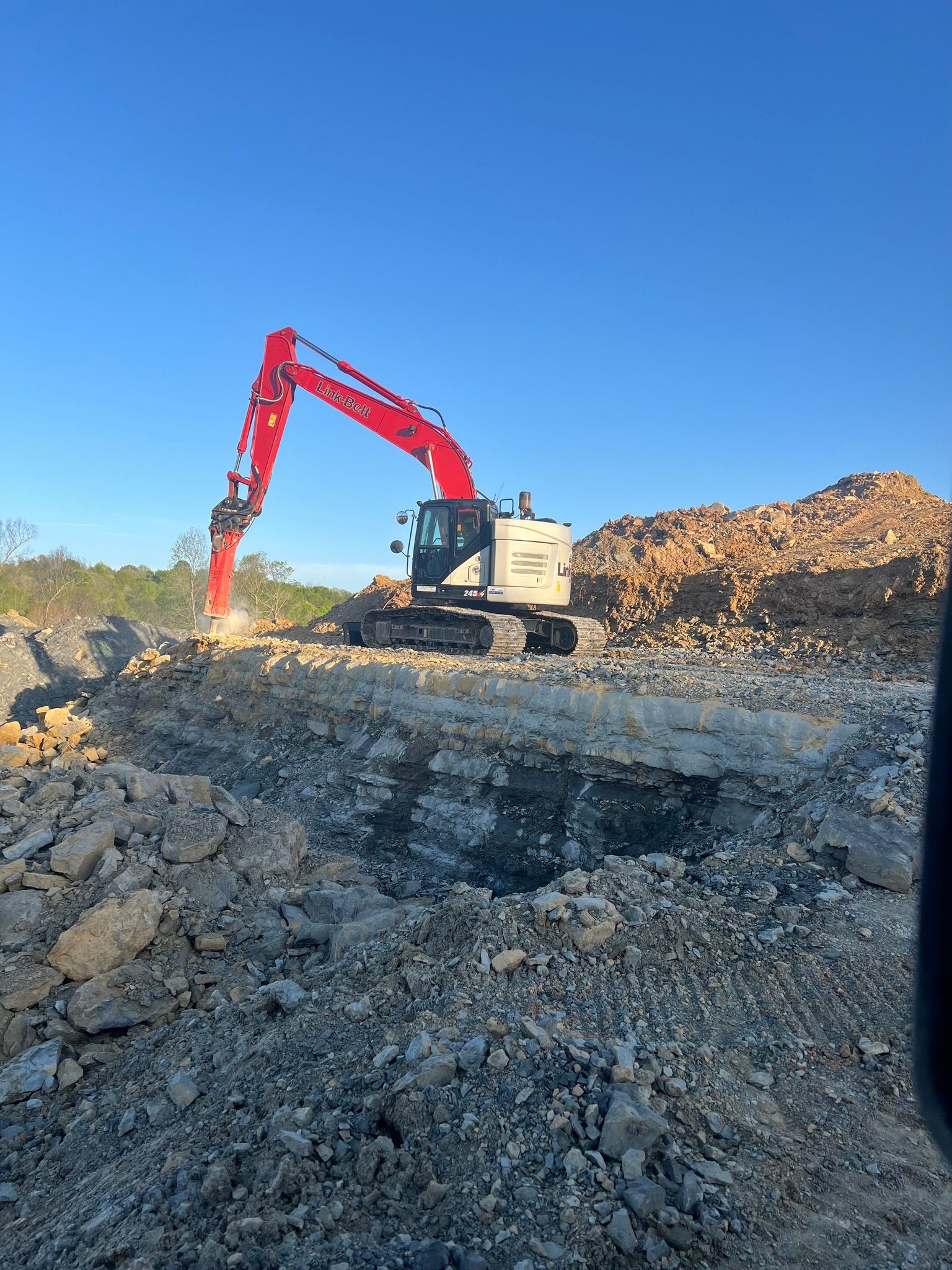 A red-and-white excavator works on a large pile of rocks and dirt under a clear blue sky.