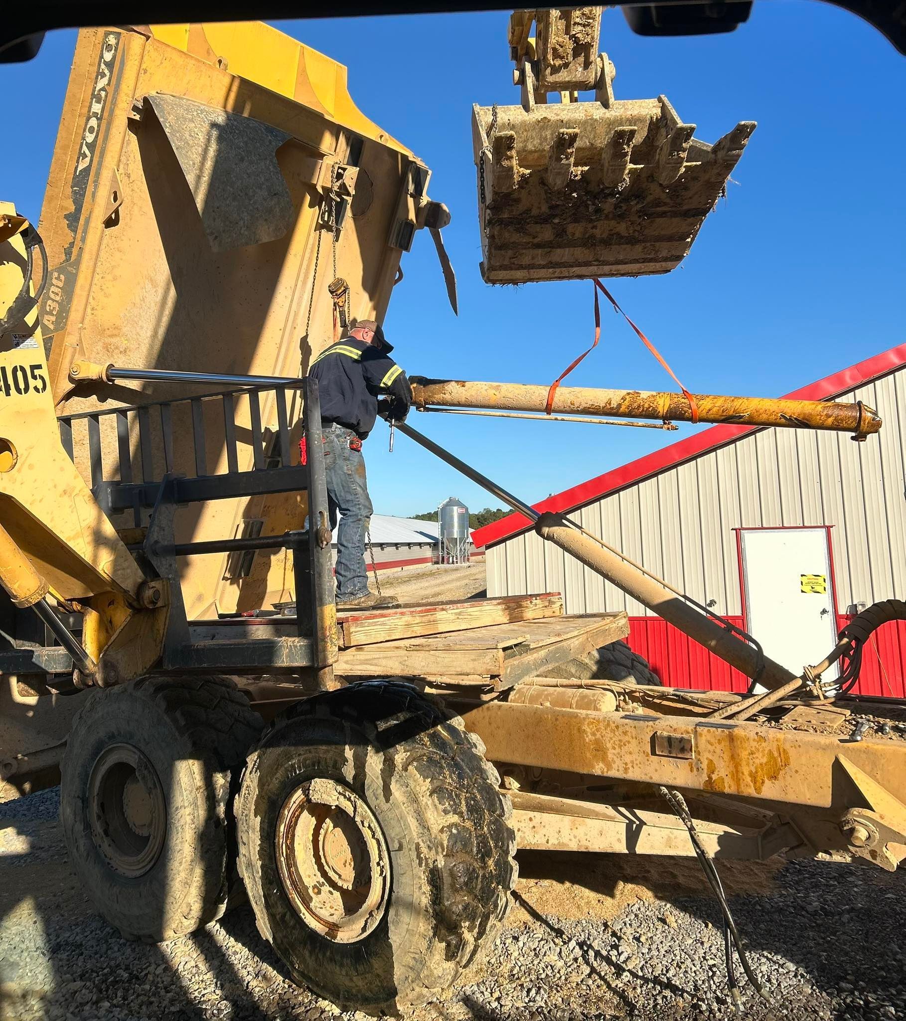 A worker wearing a hard hat guides a suspended pipe being lowered by an excavator into the bed of a yellow dump truck.