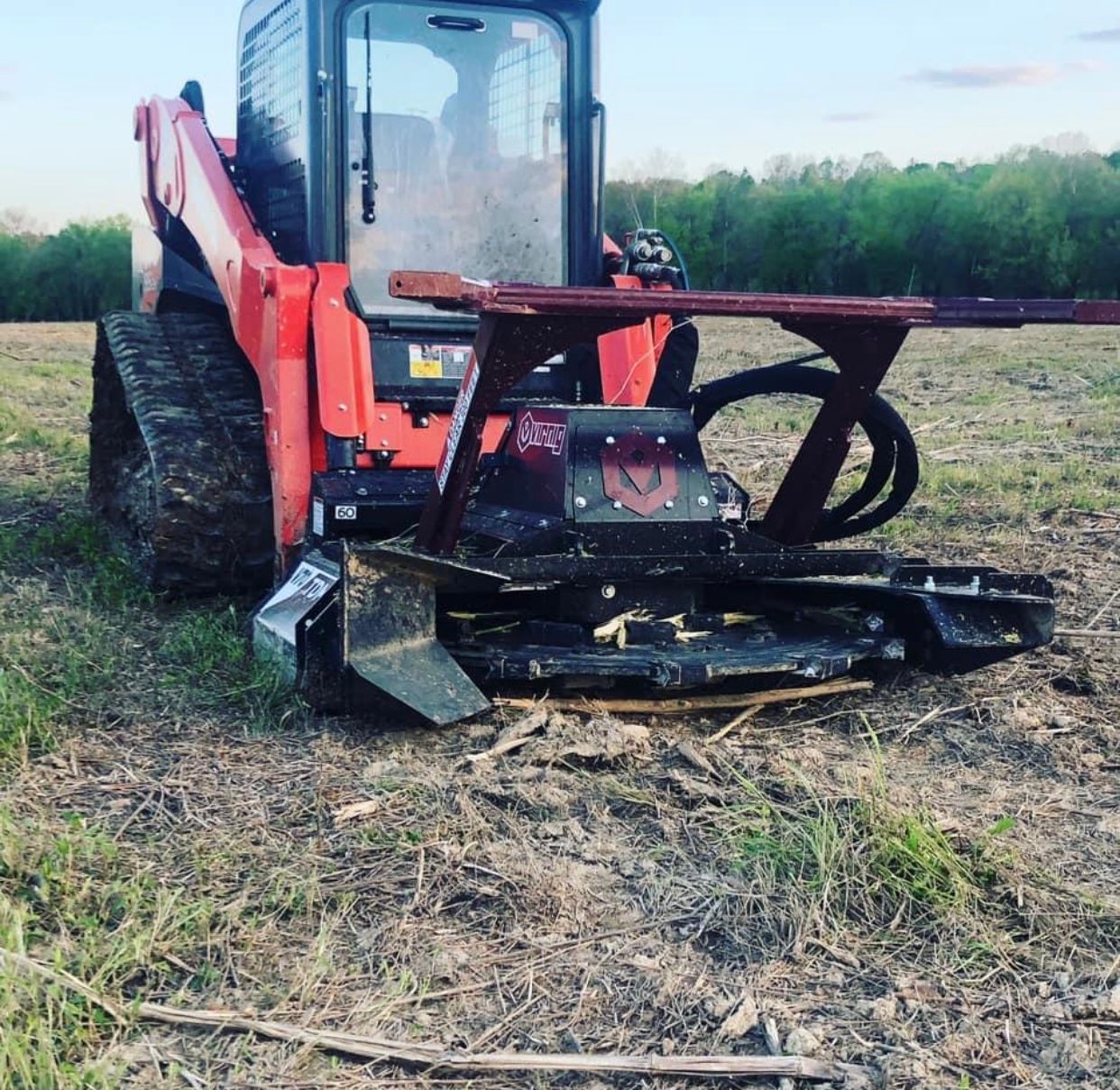 A red tracked skid steer loader equipped with a heavy-duty brush cutter attachment sits on a grassy, cleared field.