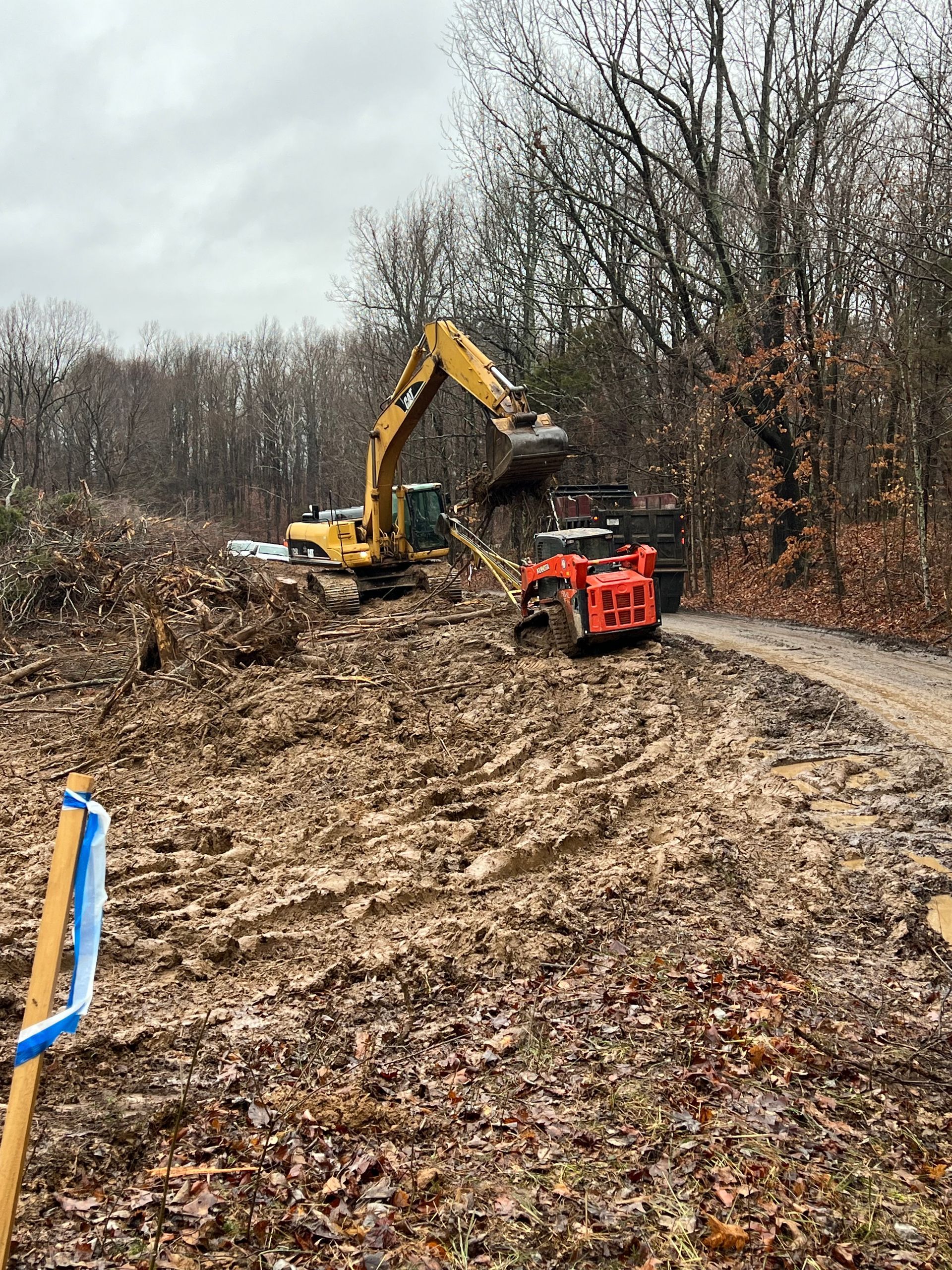 A yellow excavator loads debris into a small orange dump truck on a muddy, wooded construction site.
