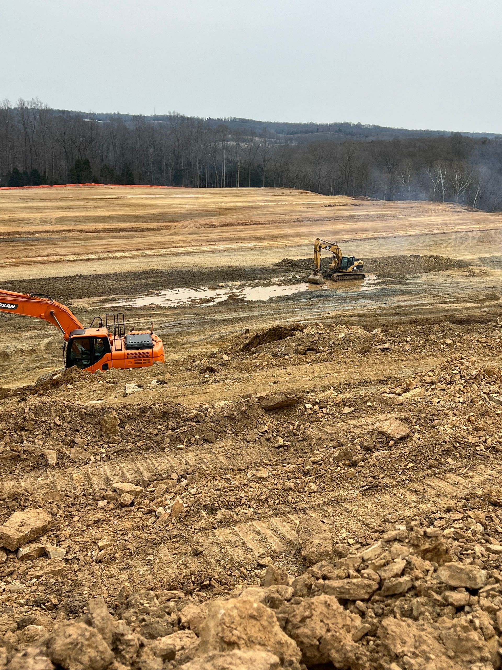 Two orange excavators operate on a muddy construction site with cleared land and trees in the distance.