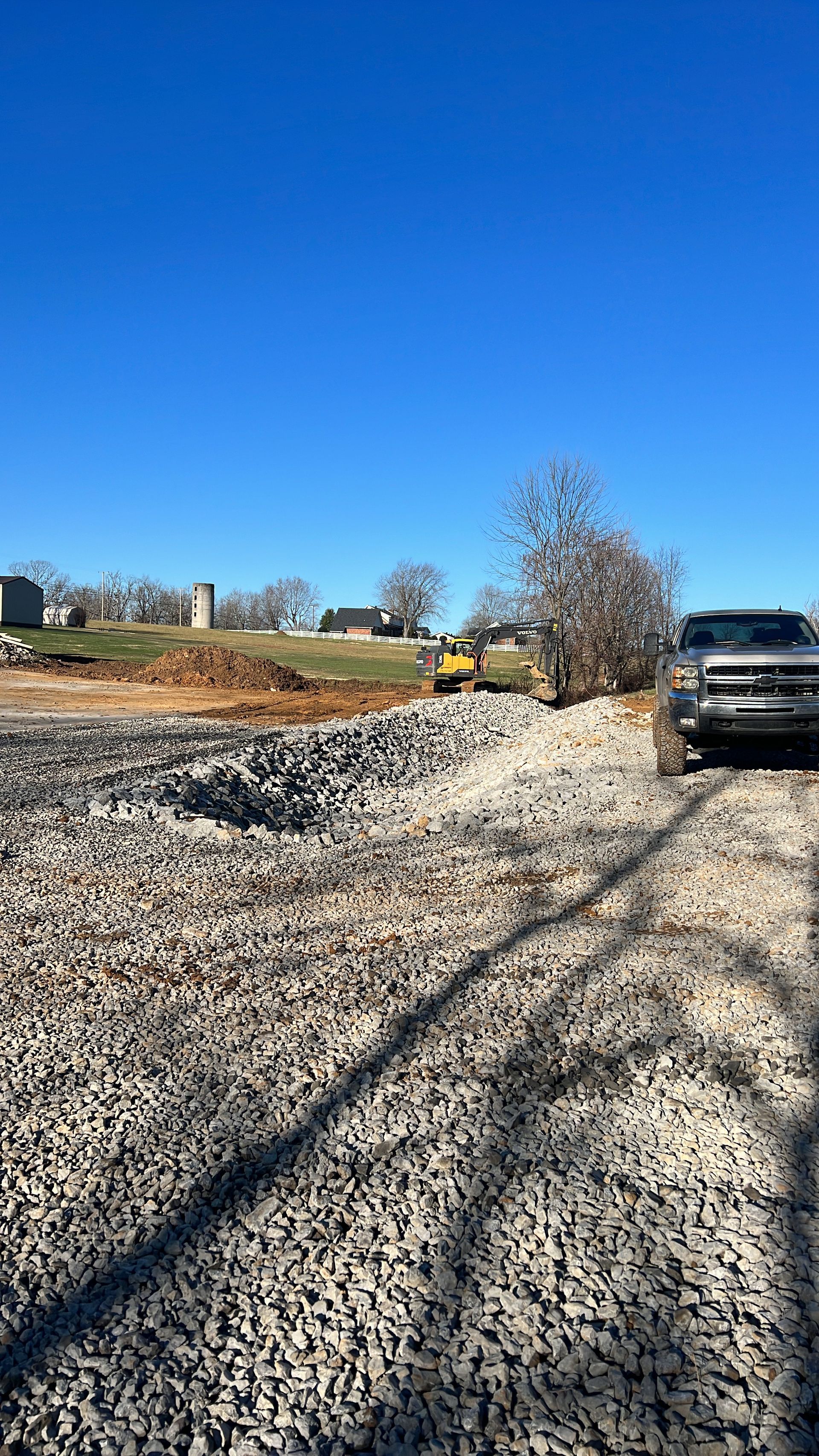 A gray pickup truck sits on a gravel construction site, with a yellow piece of heavy machinery in the distance.