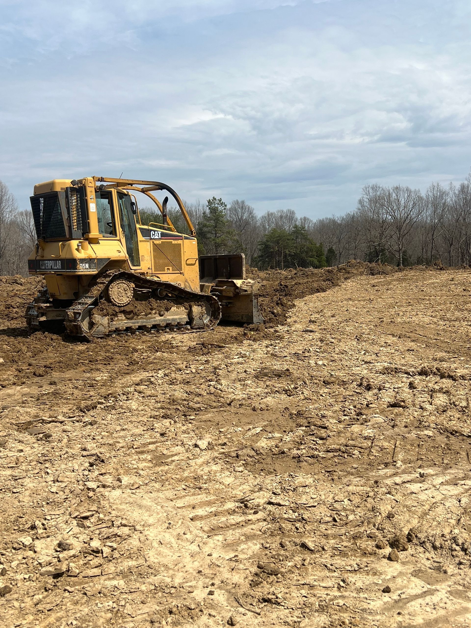 A yellow bulldozer sits on a rough, muddy, and uneven cleared field under a cloudy sky.