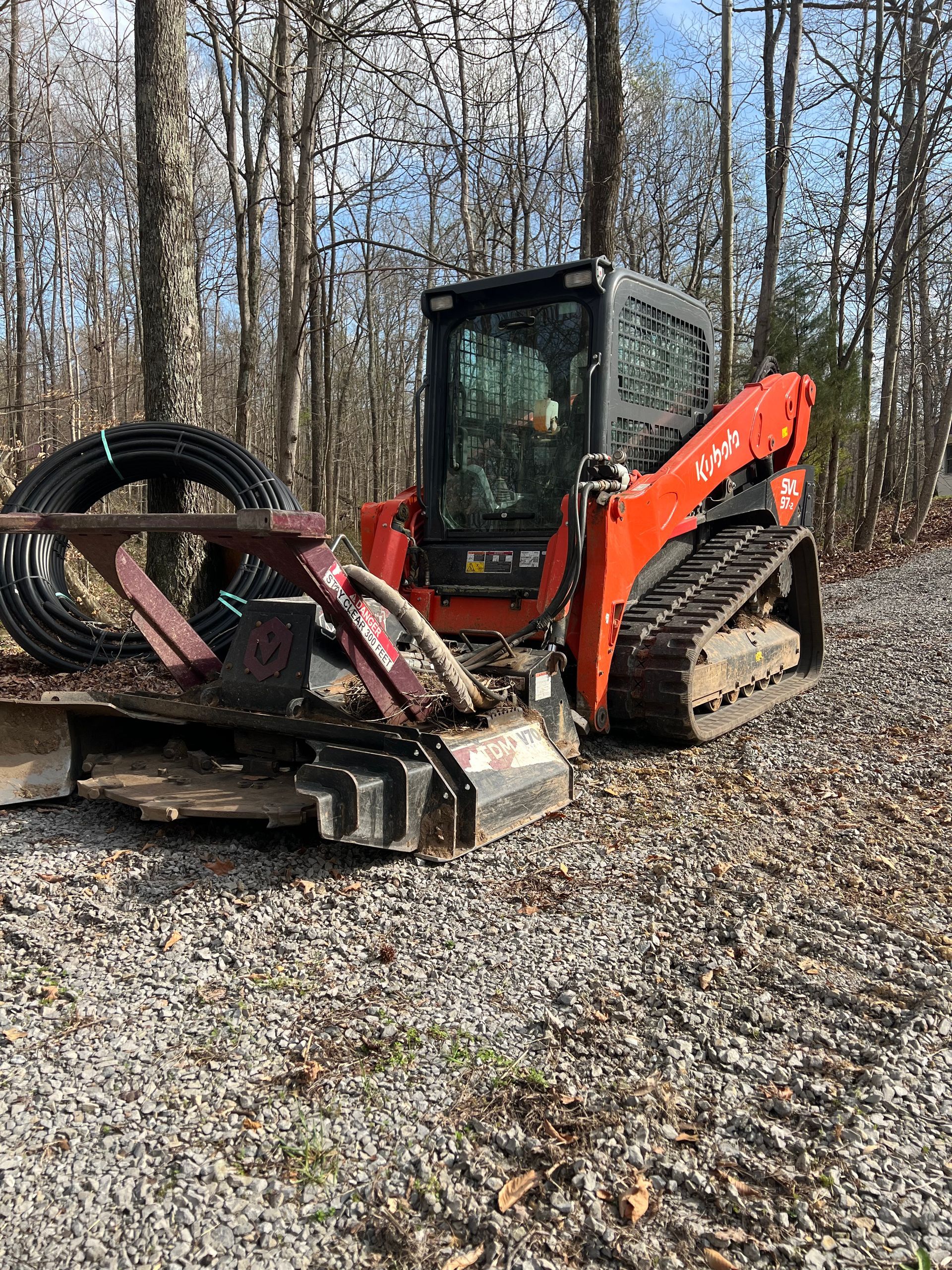 Orange Kubota skid steer with a forestry mulcher attachment parked on a gravel path near a wooded area.