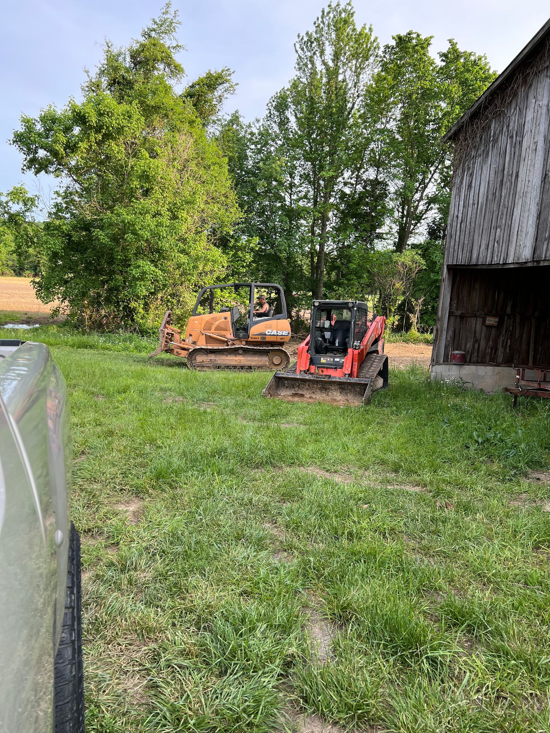 Two pieces of construction equipment, a tan bulldozer and a red skid steer, sit in a grassy field beside an old barn.