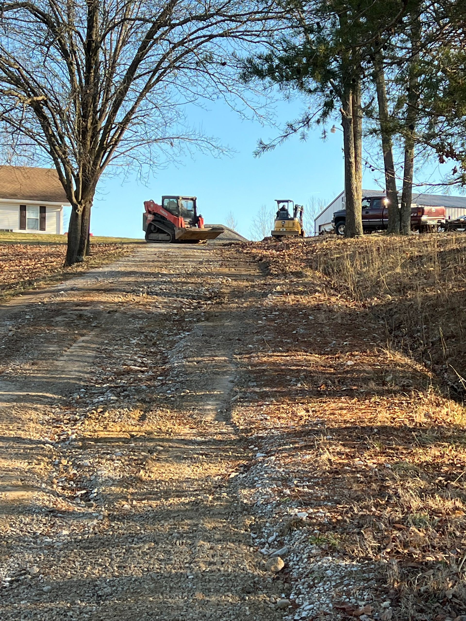 Construction equipment, including a skid steer and roller, work on a dirt driveway leading to a house in a rural setting.