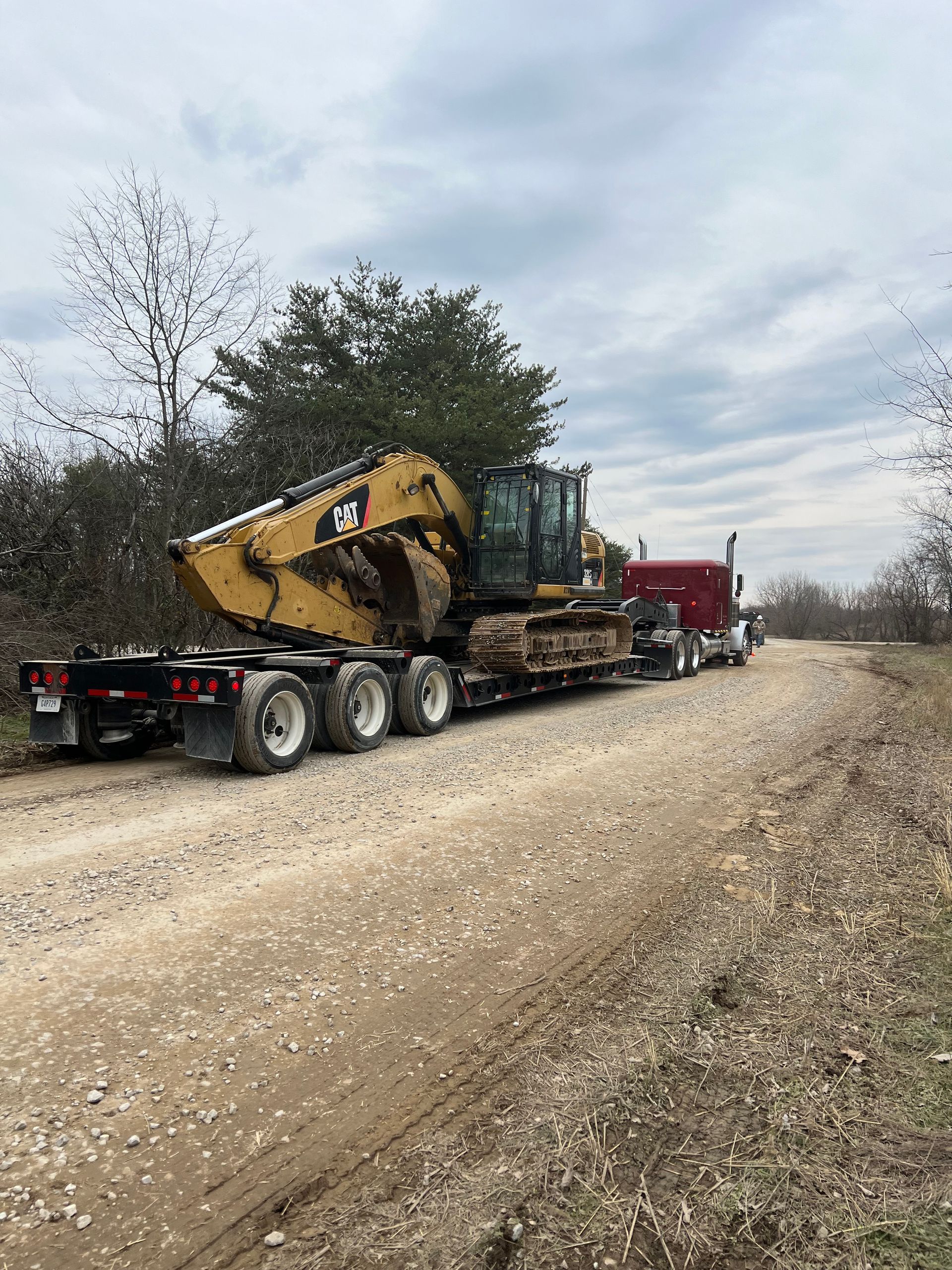 A yellow Caterpillar excavator loaded on a flatbed trailer being pulled by a red semi-truck on a gravel road.