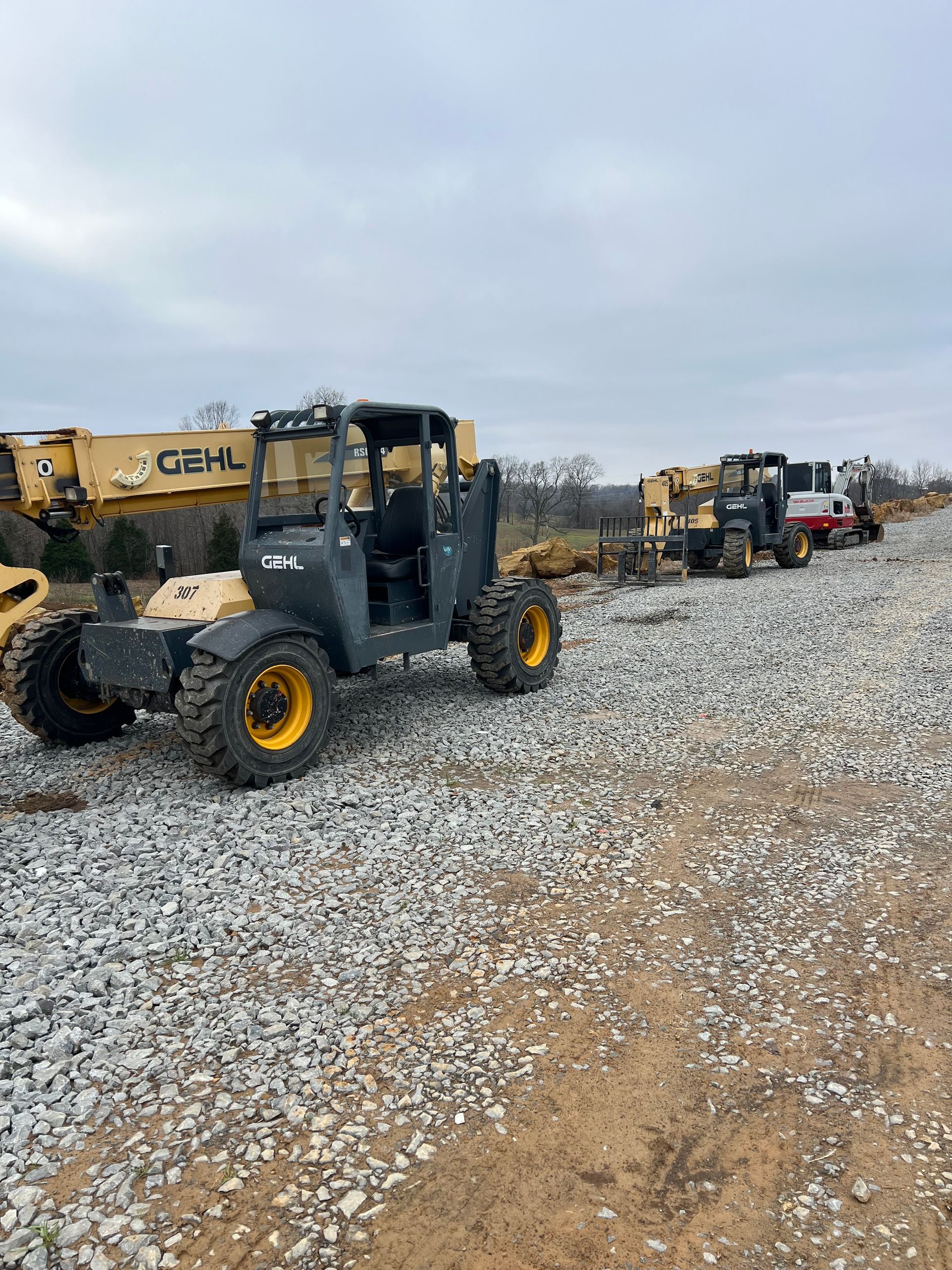 Several yellow and black heavy construction vehicles parked in a line on a gravel lot under an overcast sky.
