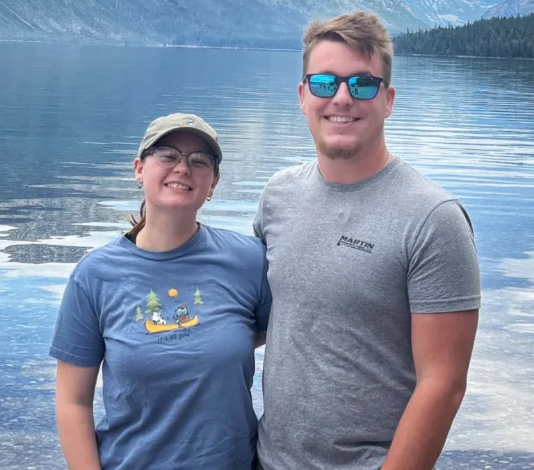 A smiling person in a cap and a person in sunglasses stand together in front of a blue lake and mountains.