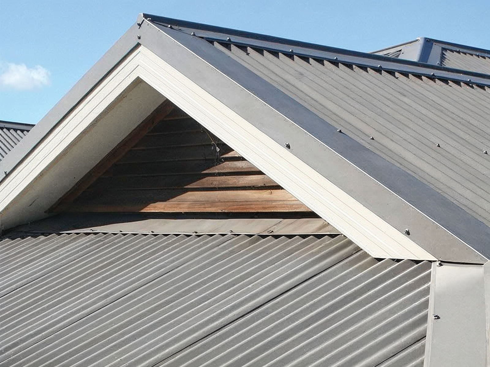 Triangular roof dormer with corrugated metal siding and white trim under a blue sky
