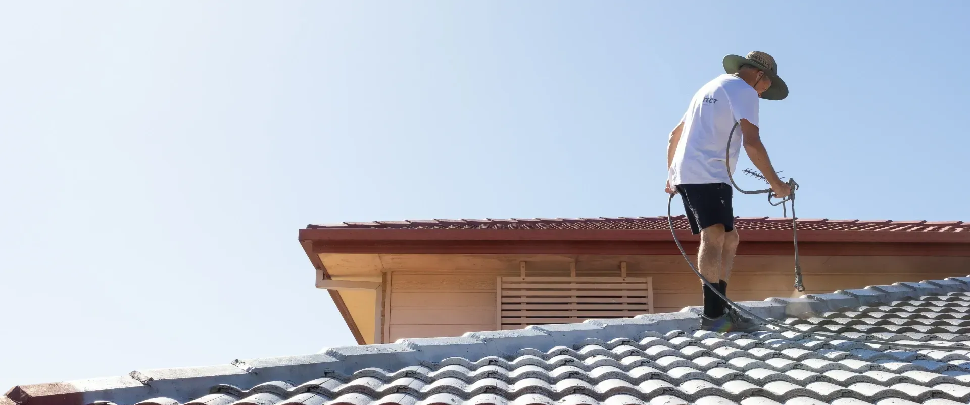Person on a rooftop inspecting tiles beside a house under a clear blue sky