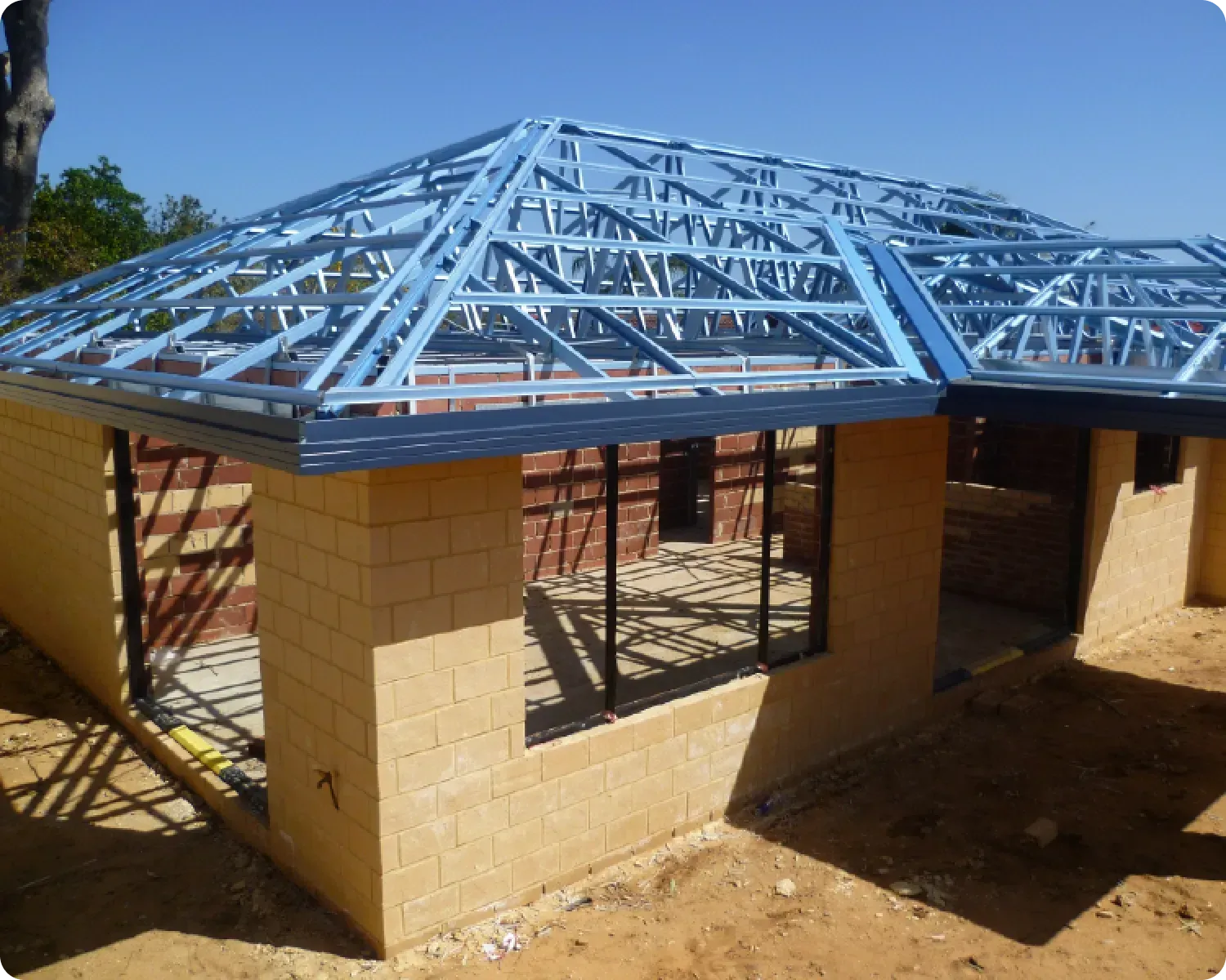 Partially built house with tan brick walls and a blue steel roof frame on a dirt lot