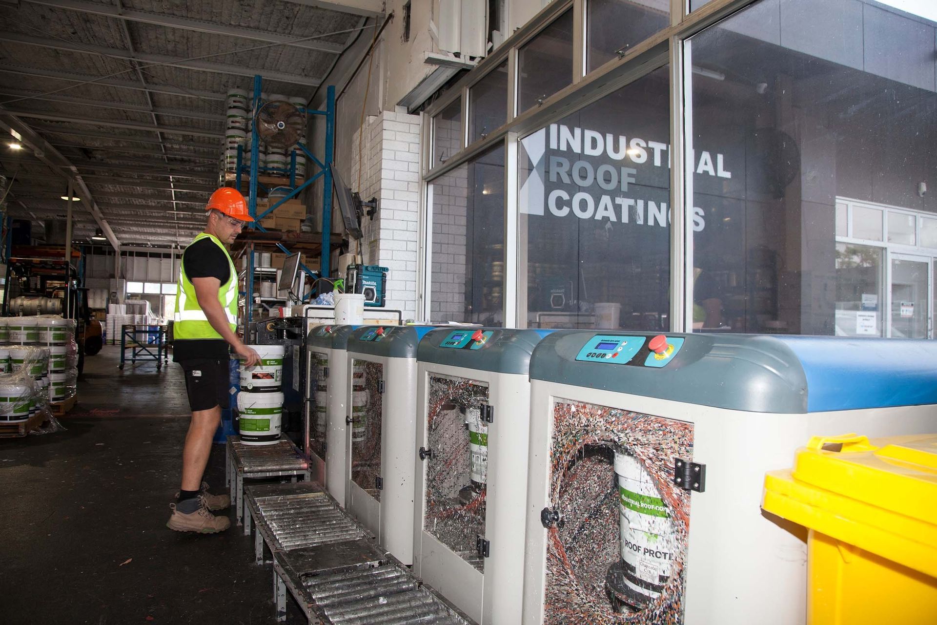 Worker in orange hard hat and black shirt stands by industrial waste bins inside a warehouse.