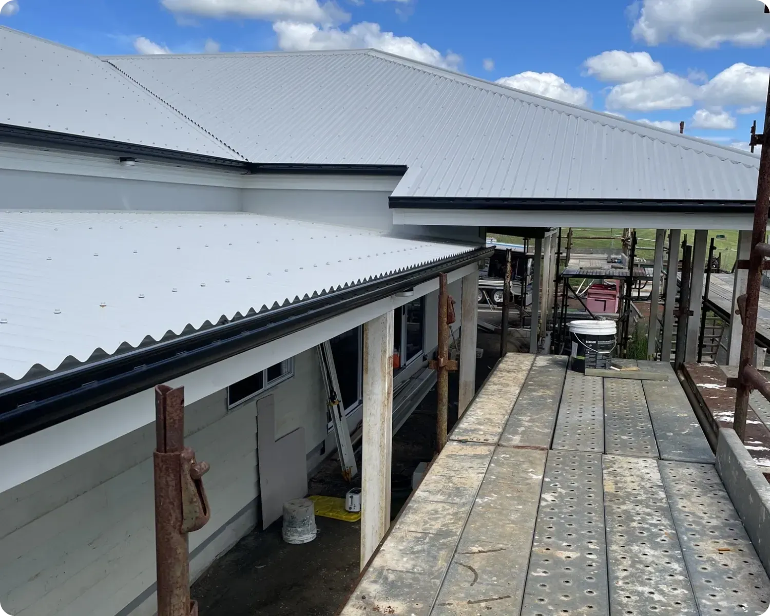 Roofed outdoor walkway beside a building, with white corrugated roofs, wooden posts, and blue sky above.