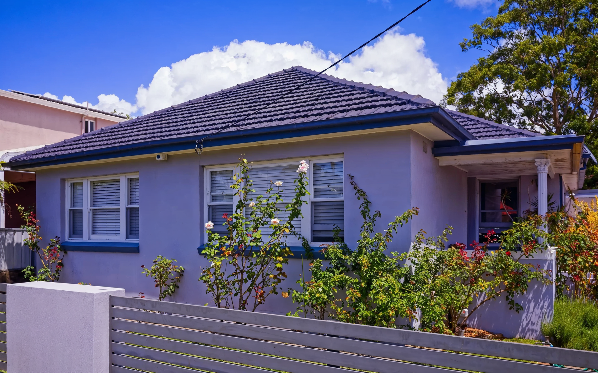 Single-story lavender house with dark tiled roof and front garden behind a low white fence.