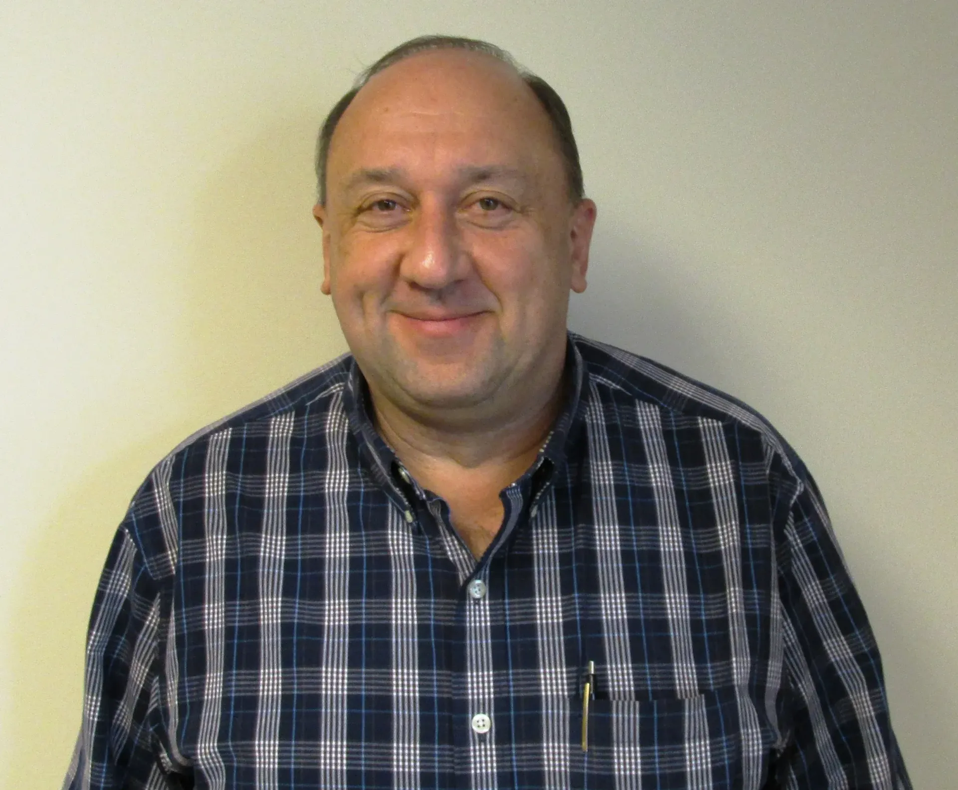Man standing against a plain wall, wearing a blue plaid shirt and smiling gently.