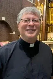 Person in clerical collar smiling indoors beside a brick wall and gold-lit altar.