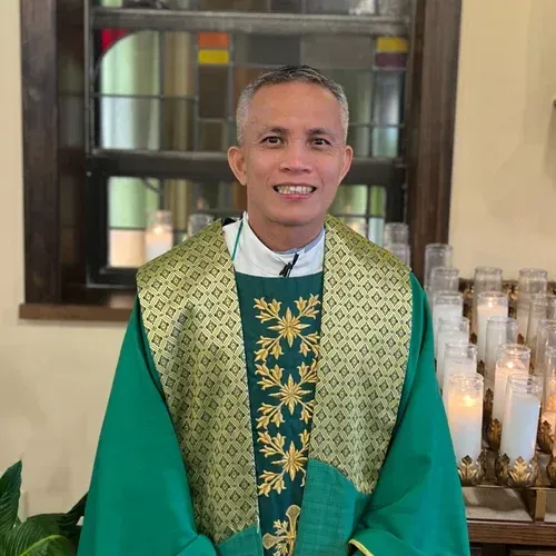 Priest in green vestments standing in a church, with candles and stained glass behind him