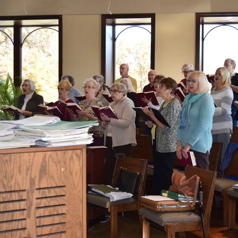 Choir members holding songbooks in a church, singing near large windows and a wooden organ.