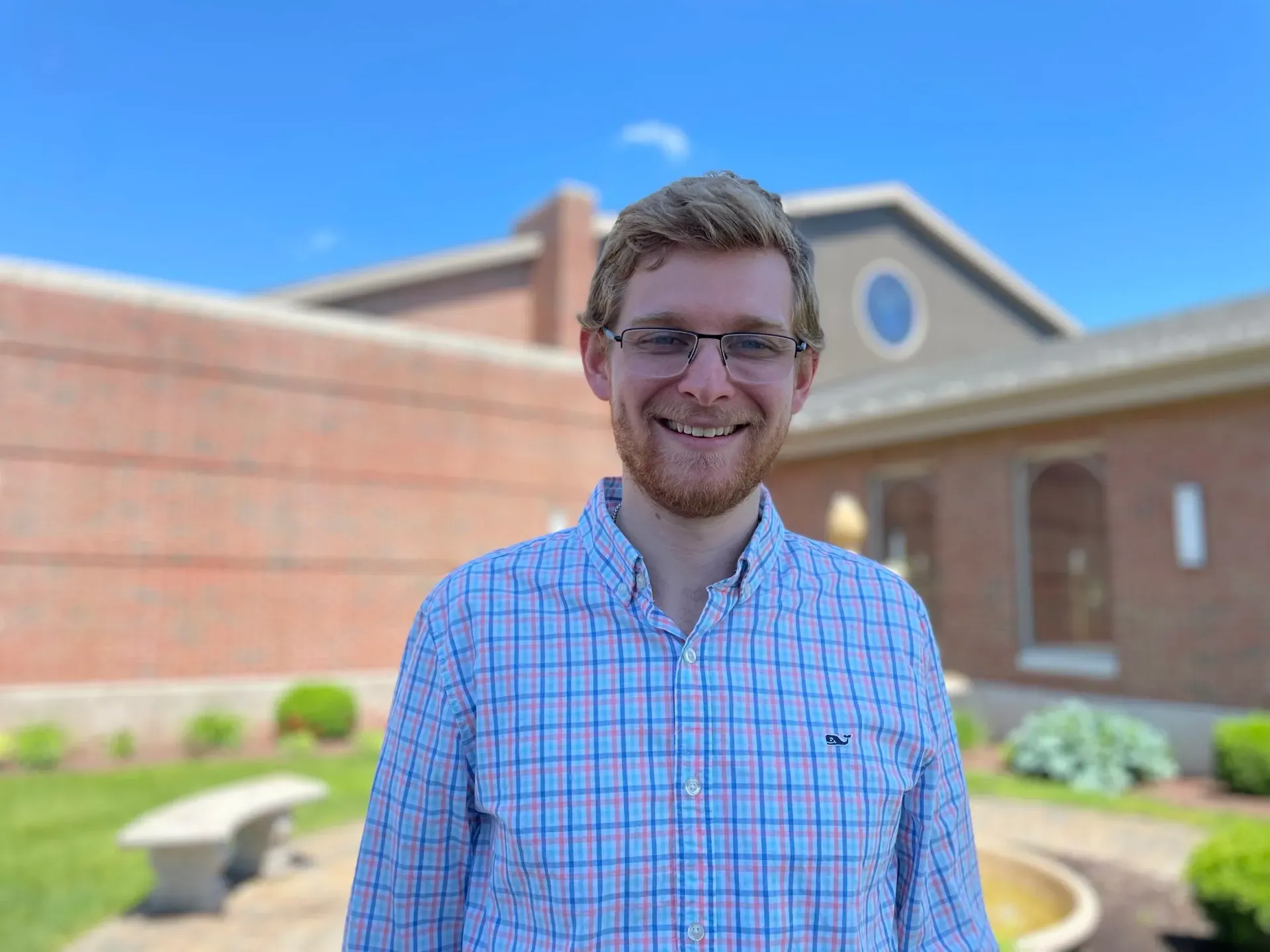 Smiling person in a blue plaid shirt standing outside a brick building on a sunny day