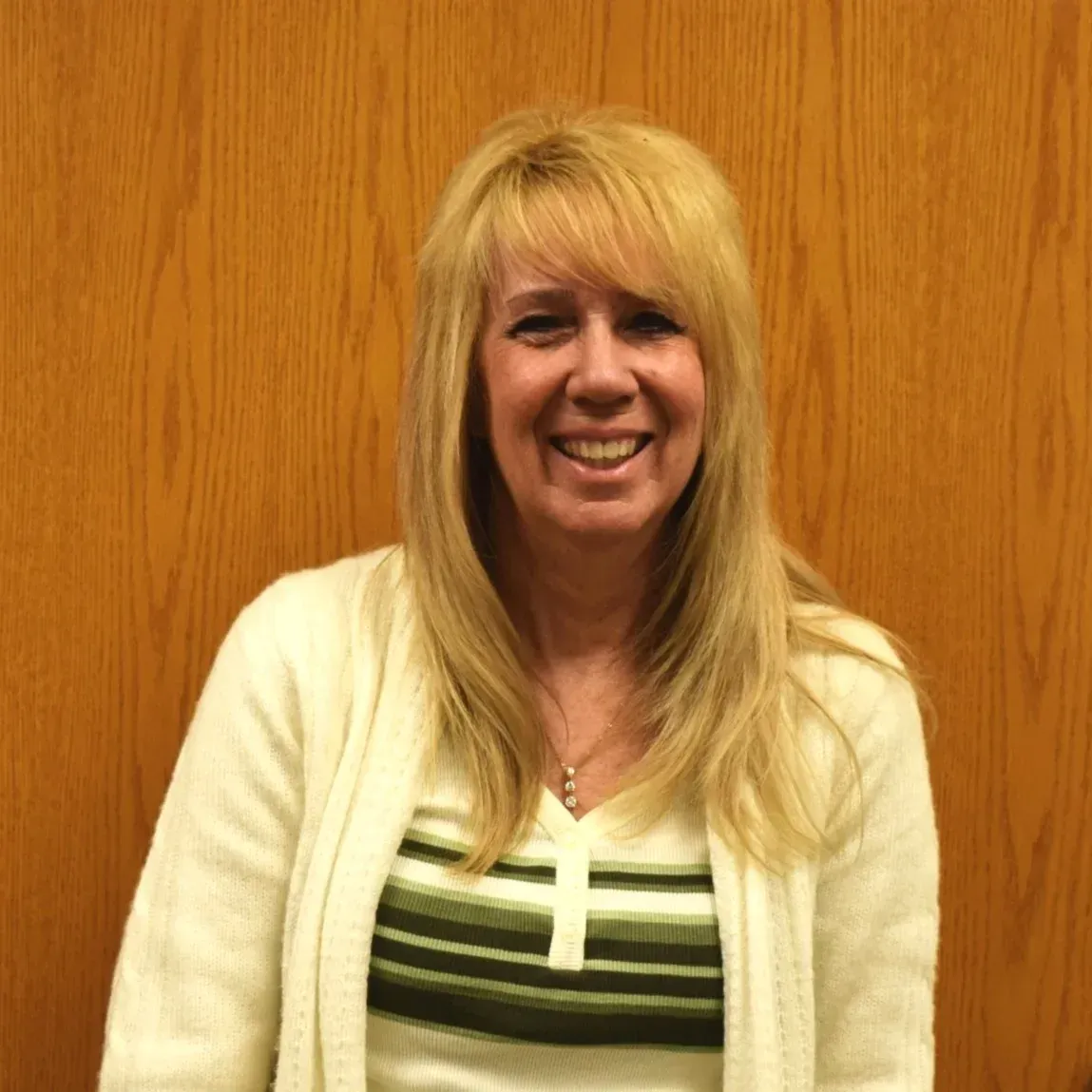 Smiling woman with long blonde hair standing against a wooden wall in a white and striped top