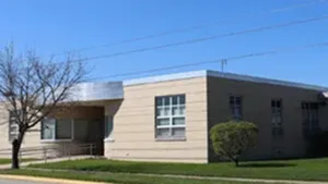 Single-story beige building with large windows and a small covered entrance beside a grassy lawn.