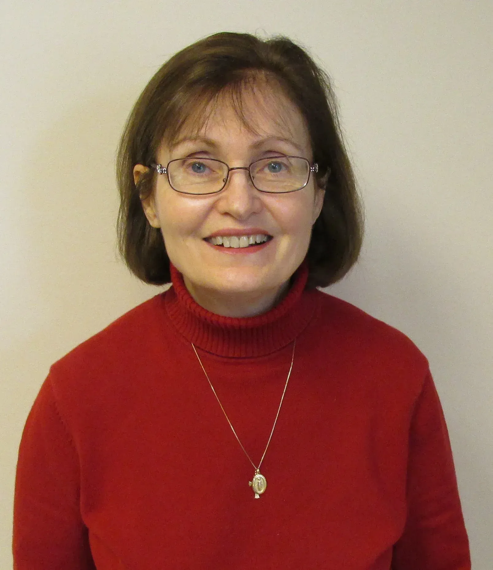 Smiling person with glasses wearing a red turtleneck and necklace against a plain background