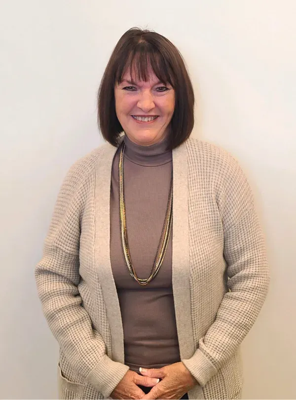 Smiling woman in beige cardigan and taupe turtleneck, standing against a plain white background.