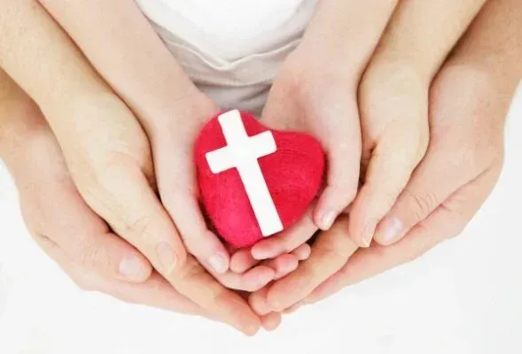 Hands cupping a red heart with a white cross on a white background