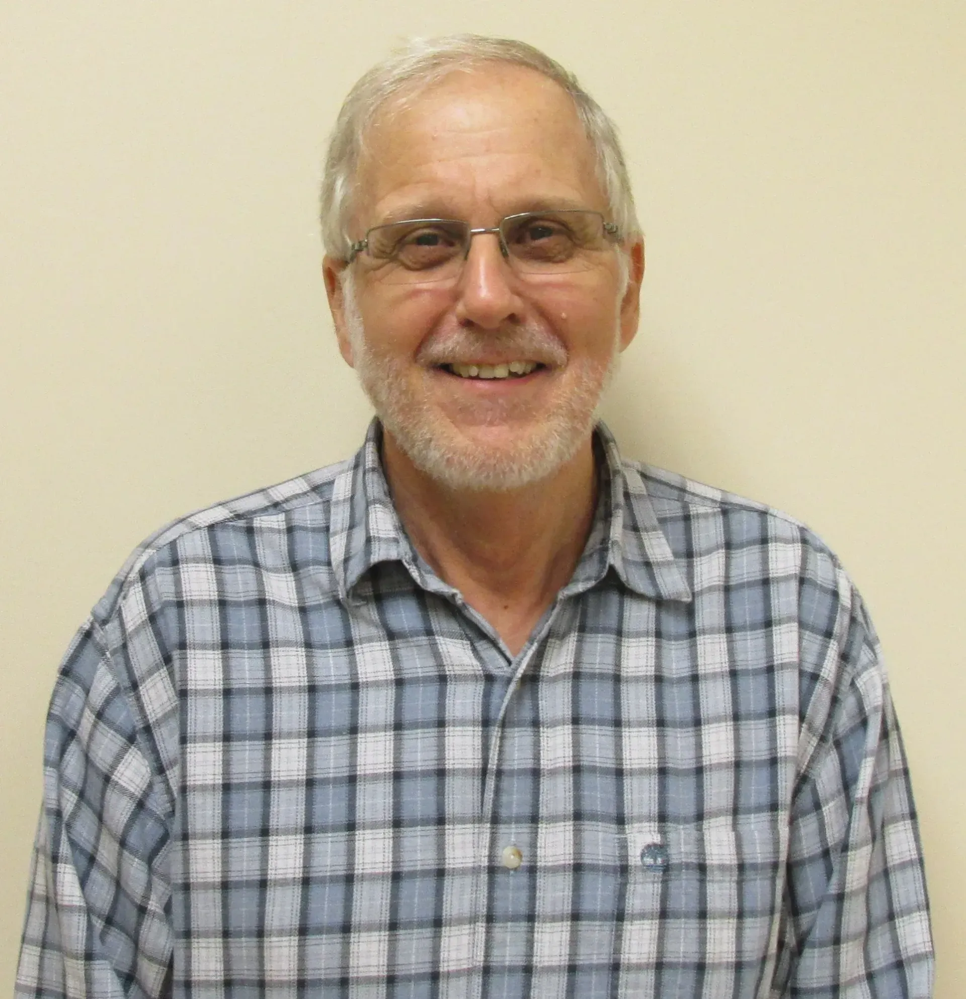 Smiling man with glasses and gray beard in a blue plaid shirt against a plain beige wall