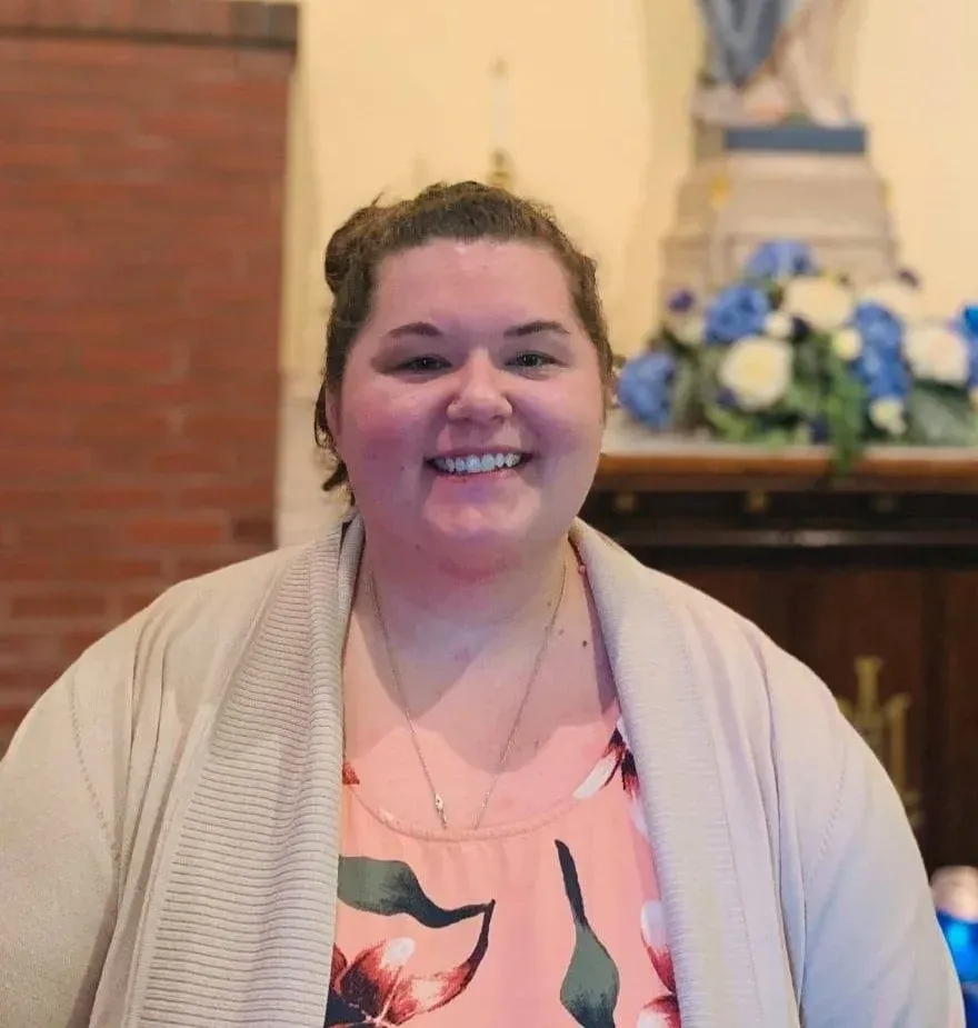 Smiling person in a peach floral top and beige cardigan, standing indoors near a decorated mantle
