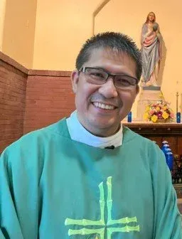 Smiling person in green clerical vestment stands inside a church near an altar and statue.