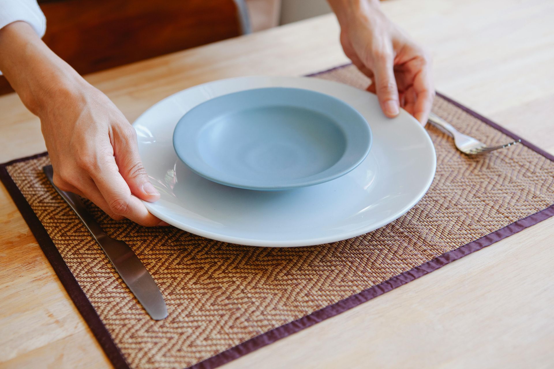 Hands placing blue bowls on a white plate over a woven placemat at a dining table