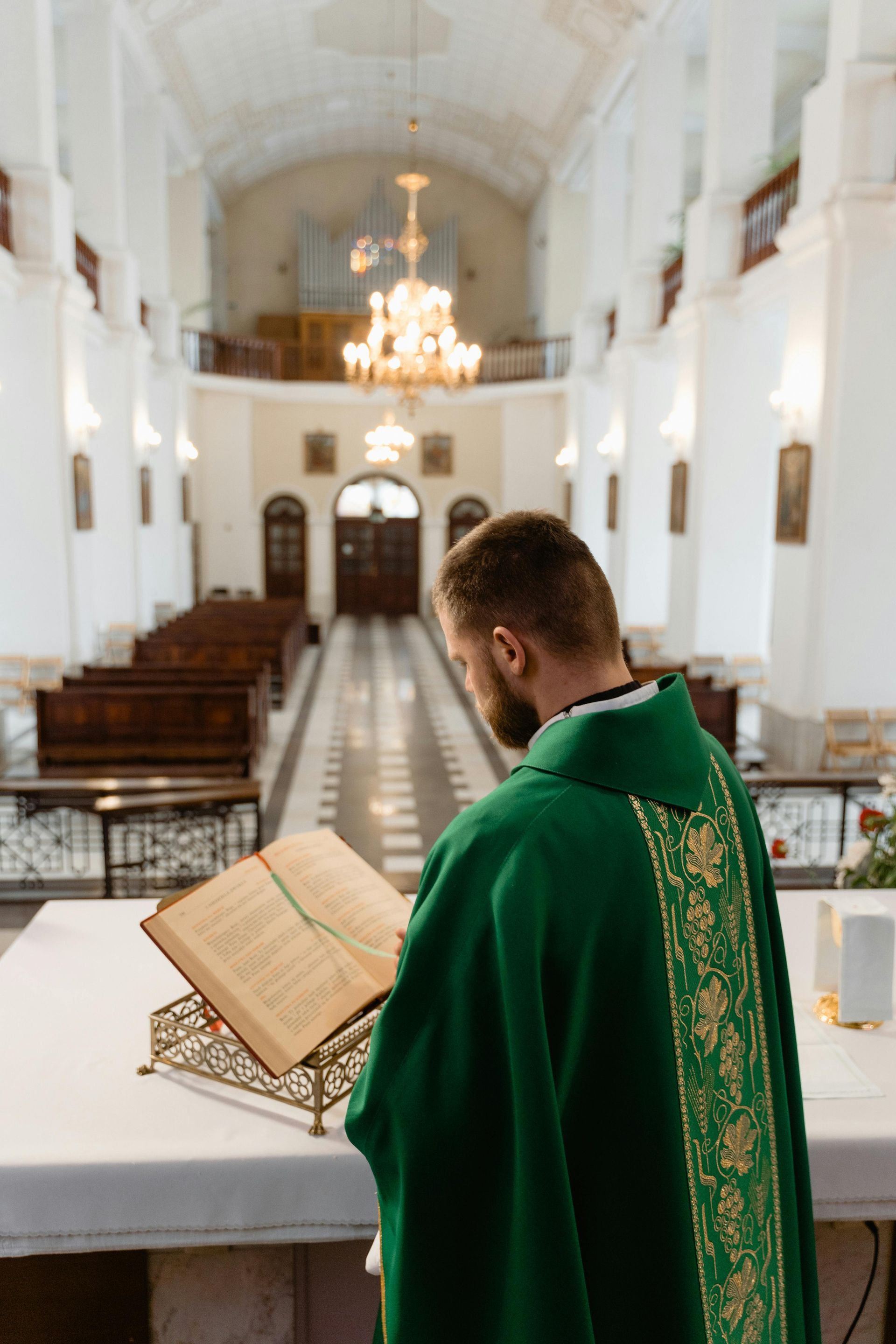 Priest in green vestments reading at an altar in a church with chandeliers and pews