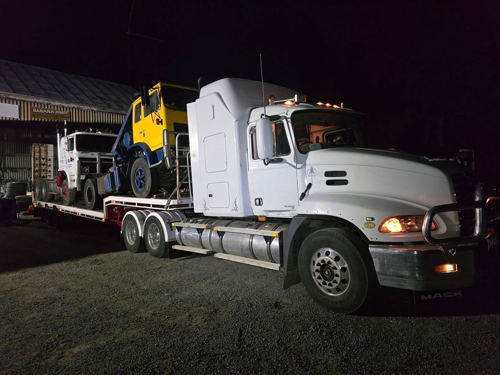 White Semi-Truck Hauling a Yellow Backhoe on A Flatbed Trailer — Rainbow and Sons Heavy Haulage in Wells Crossing, NSW