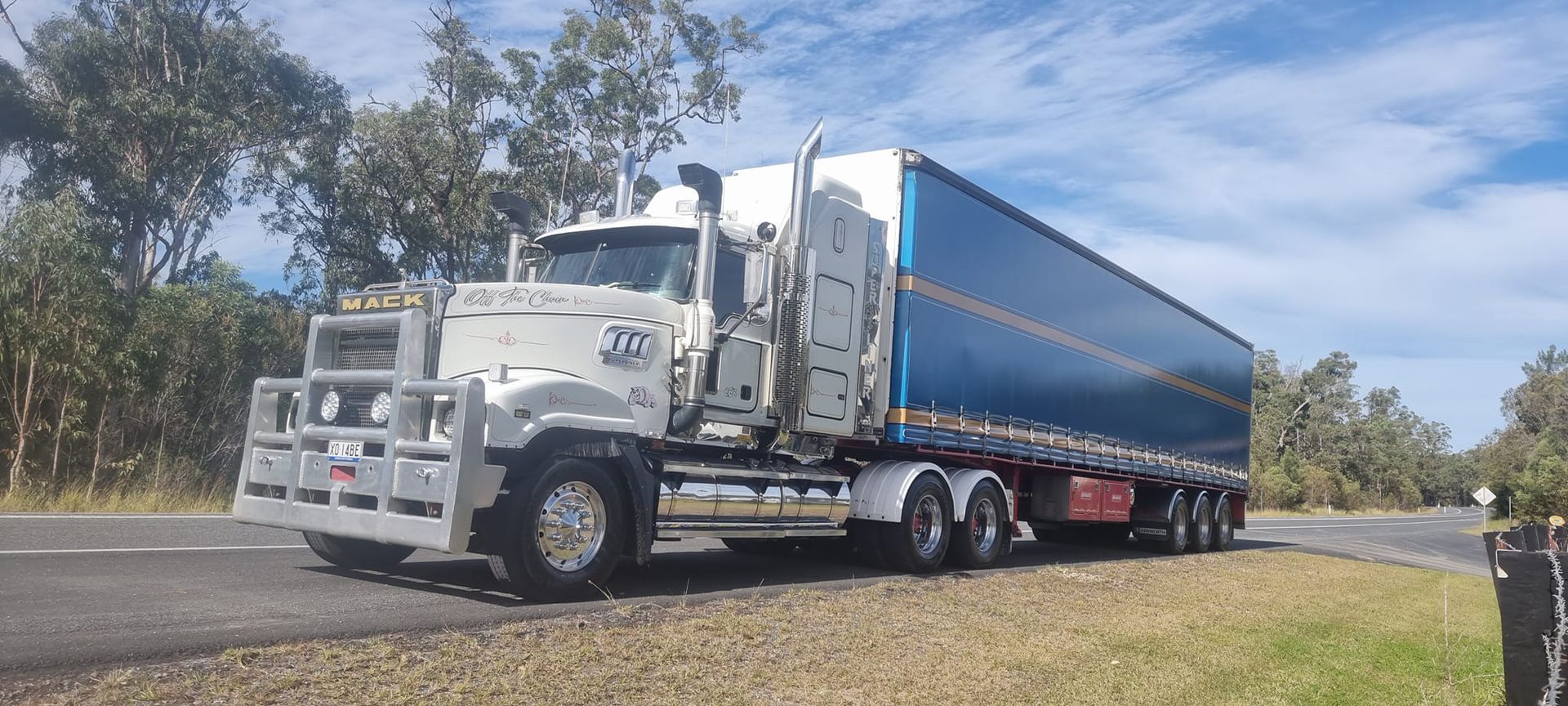 White semi-truck with blue trailer driving on a rural road under trees — Rainbow and Sons Heavy Haulage in Wells Crossing, NSW