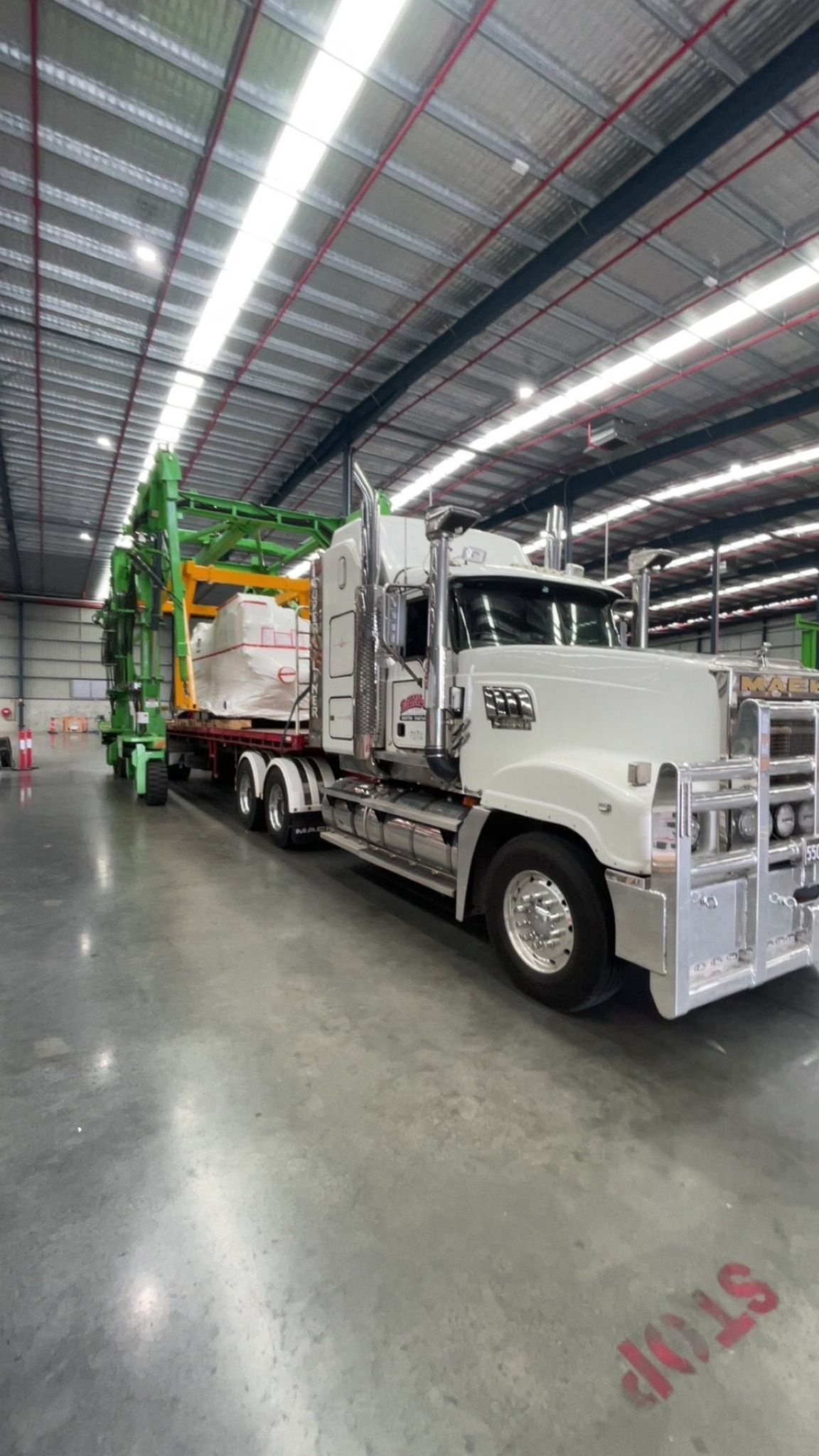 White Garbage Truck With Green and Yellow Lift Parked Inside a Large Warehouse at Night — Rainbow and Sons Heavy Haulage in Coffs Harbour, NSW