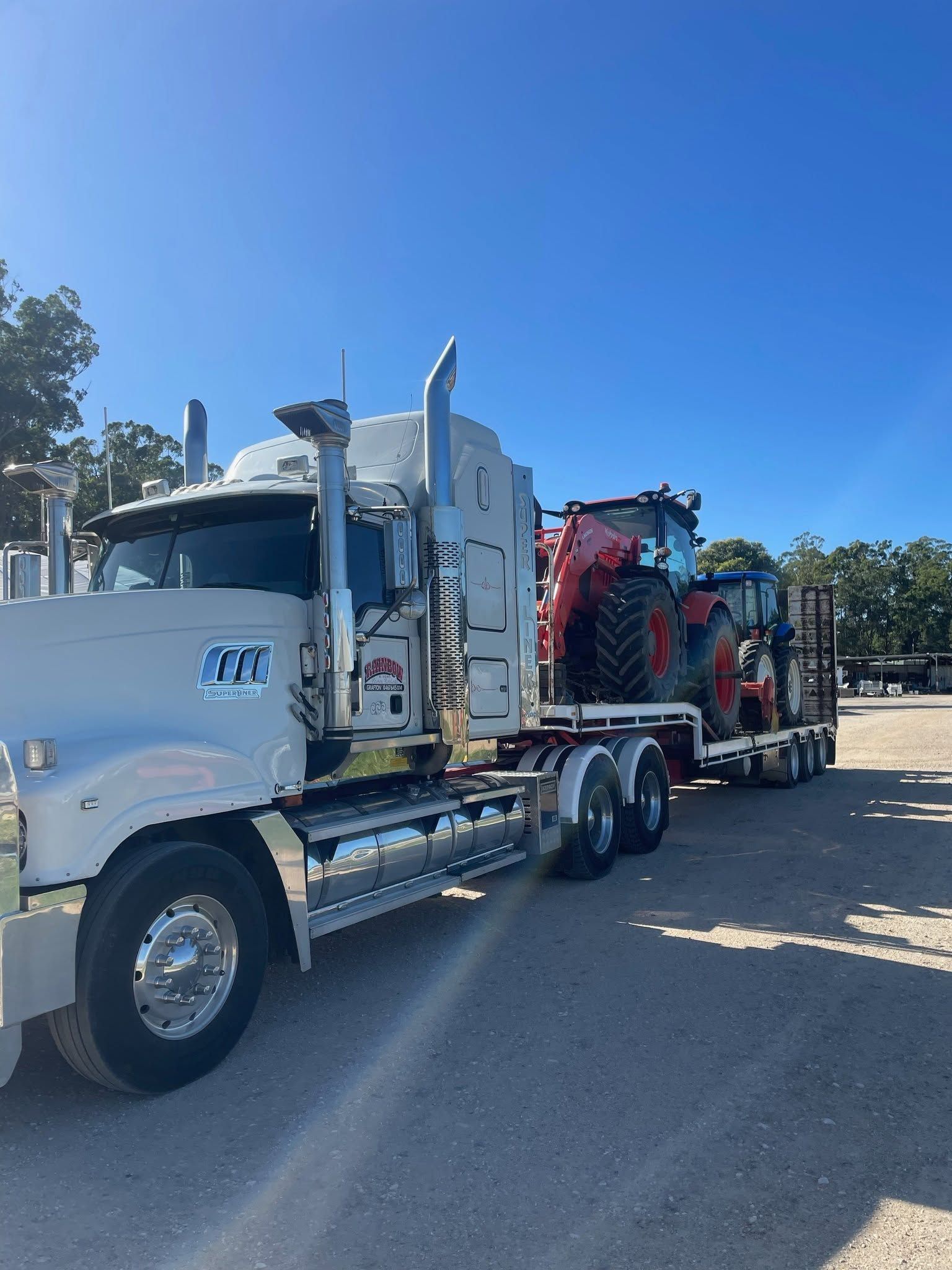 White Semi Truck Hauling Red Machinery on A Flatbed Trailer — Rainbow and Sons Heavy Haulage in Wells Crossing, NSW