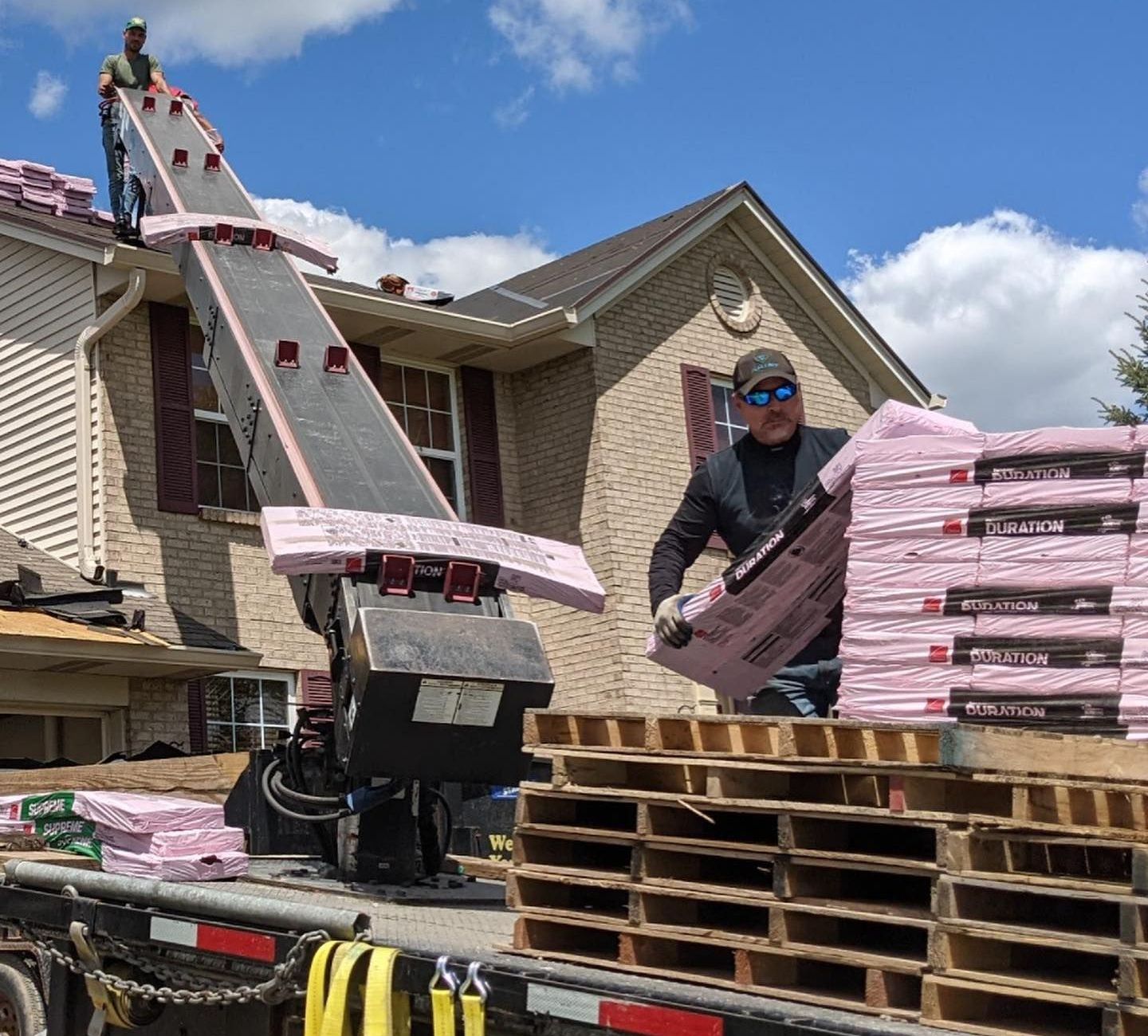 Construction worker loads pink insulation onto a roof using an angled conveyor. Truck with pallets. Blue sky.