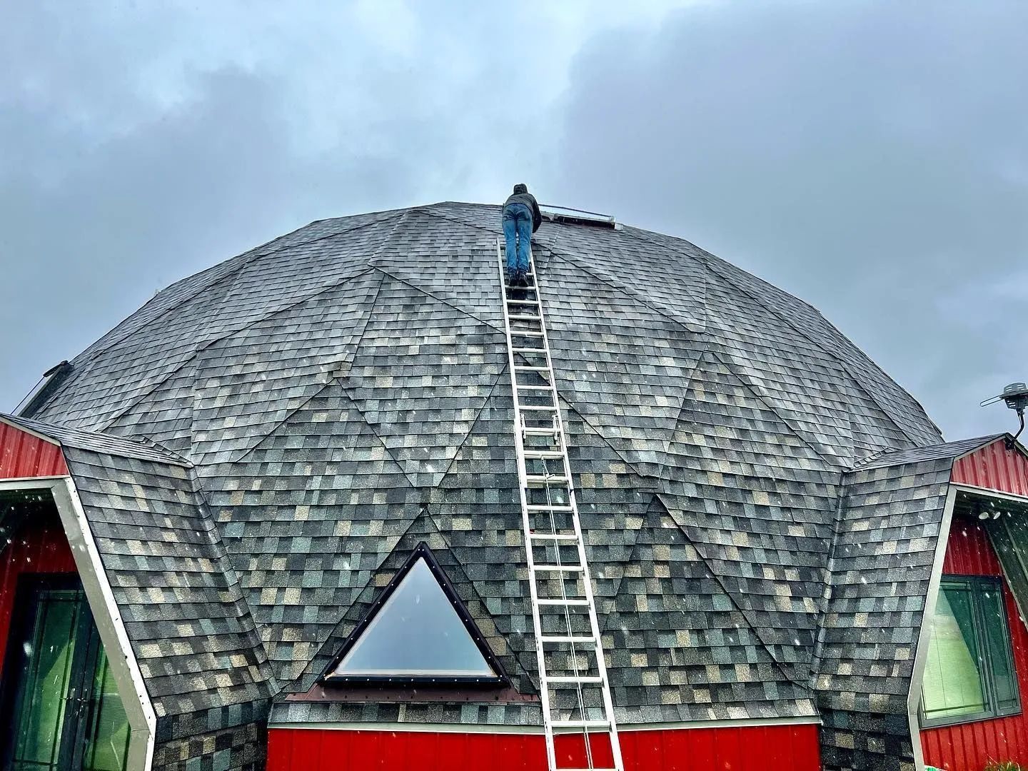 Person on ladder repairing shingles on a large, dome-shaped building under a cloudy sky.