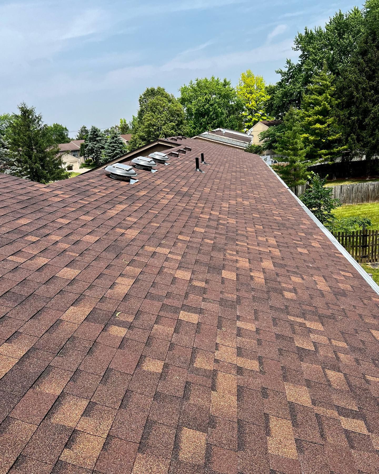 Brown shingled roof with vents, viewed from above on a sunny day with trees and blue sky in the background