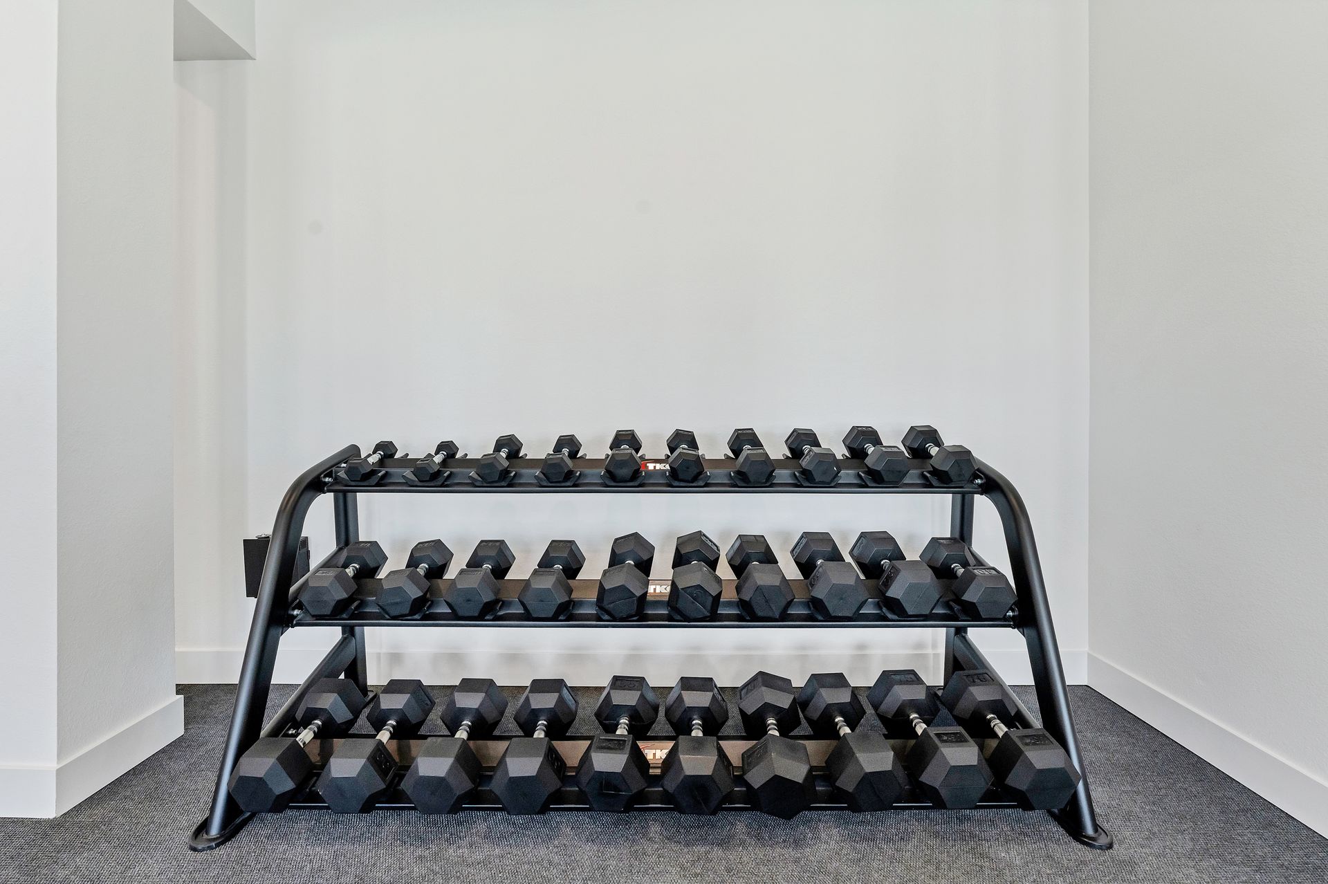 Three-tier dumbbell rack with rows of black weights in a white-walled gym room