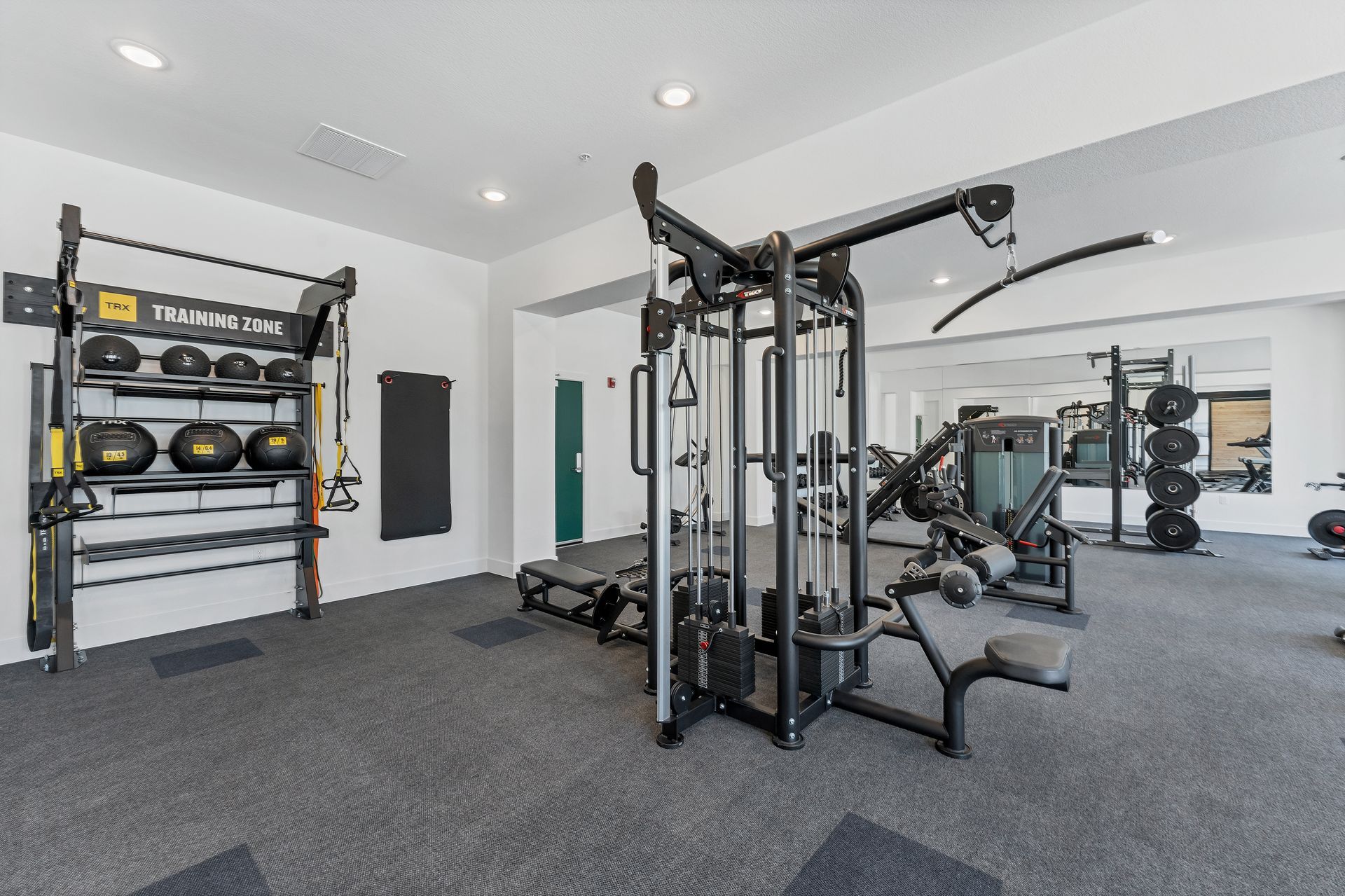 Empty gym with black exercise equipment, mirrors, and gray carpeted floor under bright ceiling lights
