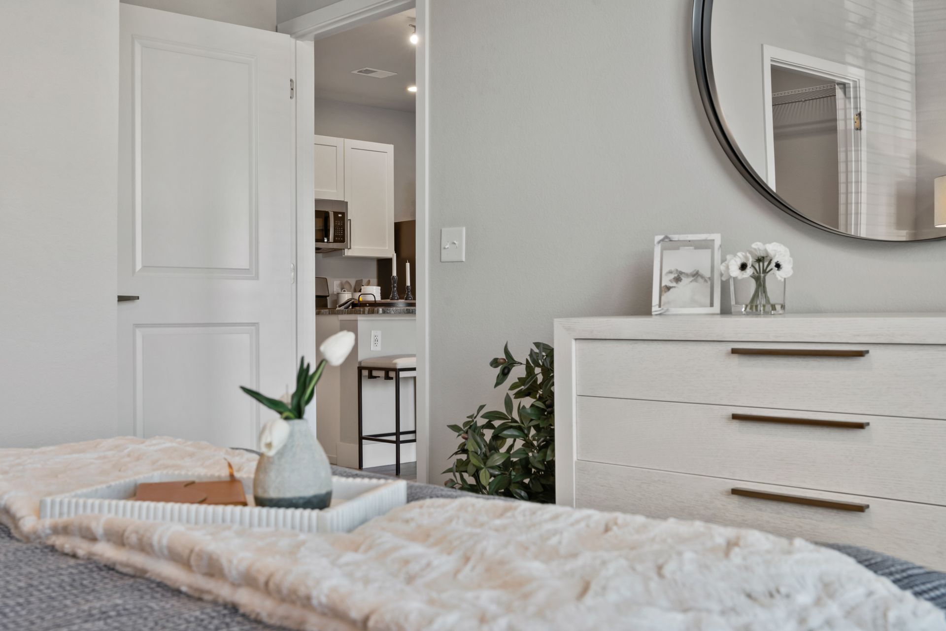 Bright bedroom with white dresser, bed, mirror, plant, and doorway to a kitchen beyond.