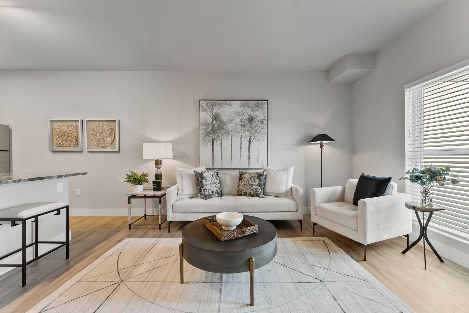 Bright modern living room with white sofa, armchairs, round coffee table, and large window blinds.