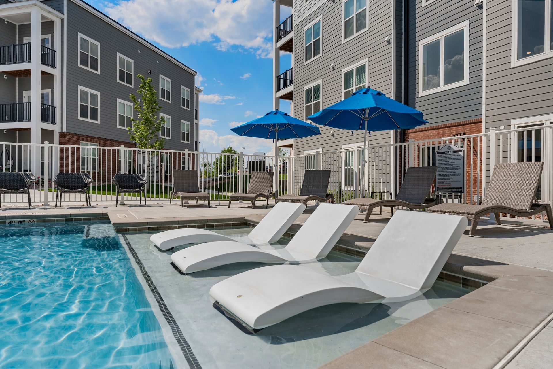 Apartment pool deck with white lounge chairs, blue umbrellas, and modern buildings around a swimming pool