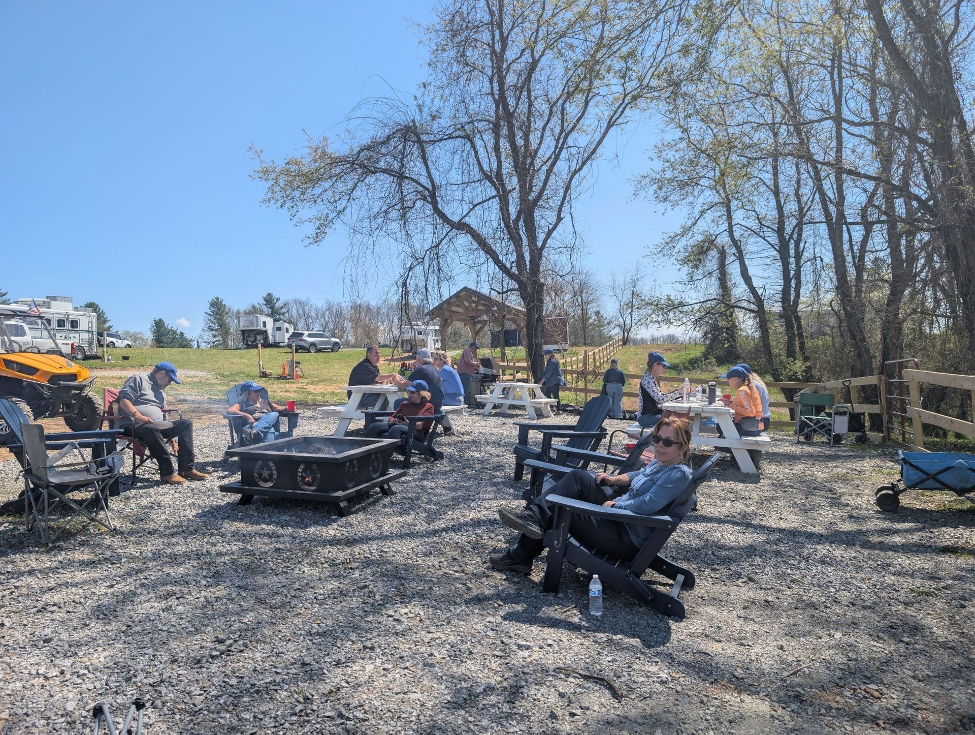 People seated outdoors around fire pits on a sunny gravel patio with leafless trees and a blue sky.
