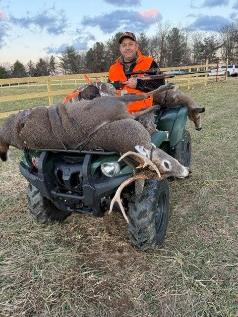 Person in orange vest on ATV with a deer carcass in a field at dusk