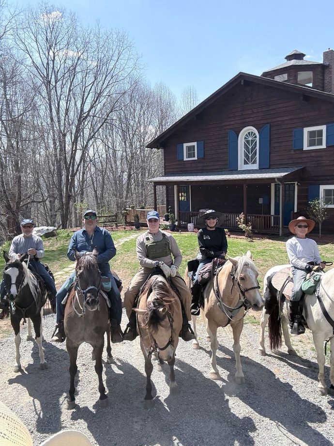 Six people riding horses in front of a brown house on a sunny day