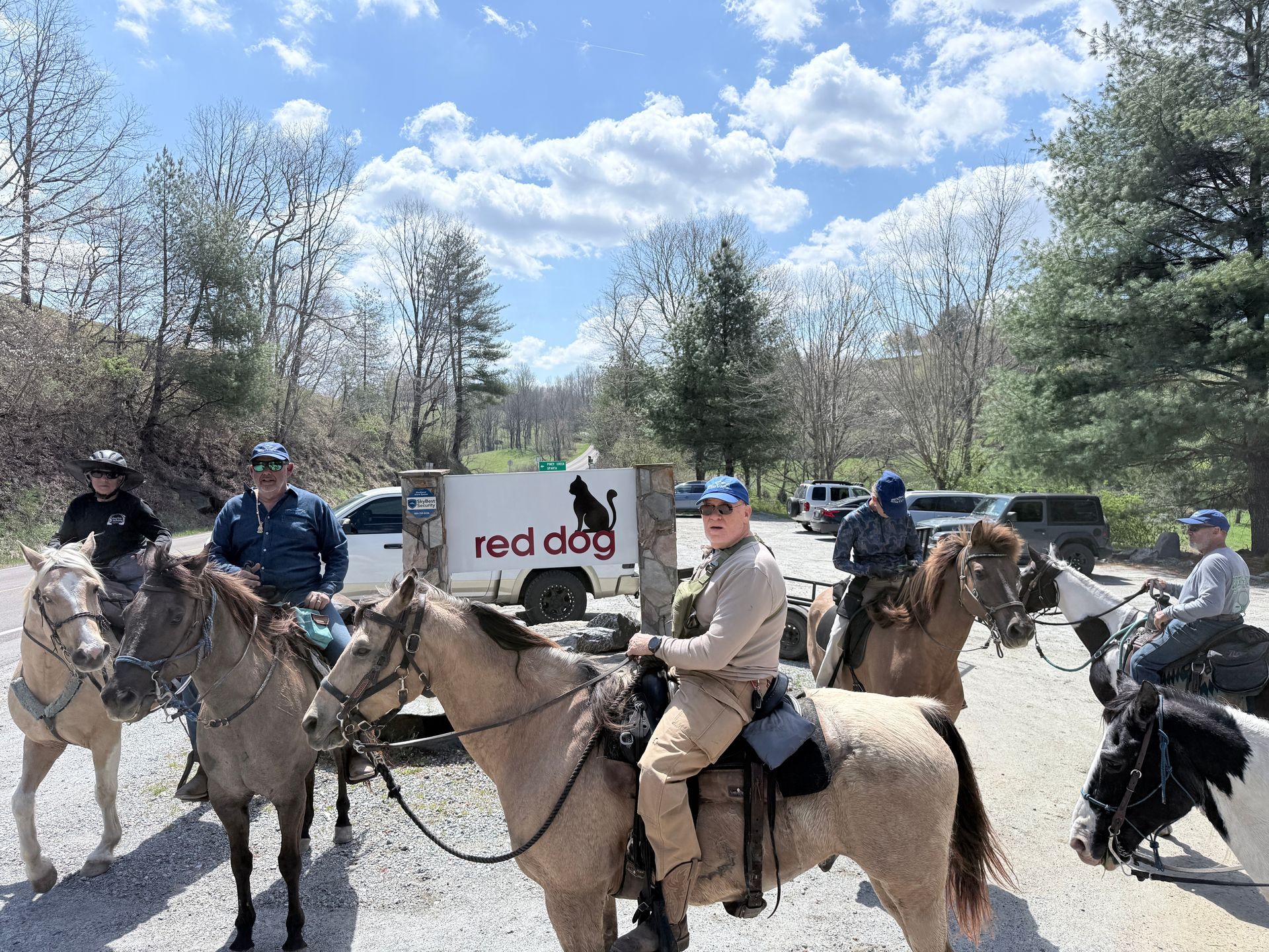 Mounted riders on horseback outdoors in front of a red dog sign on a sunny day